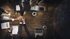 Vue aérienne d’une équipe de professionnels en costume réunis autour d’une table, posant leurs mains l’une sur l’autre en signe de collaboration, avec des documents, ordinateurs et café sur la table.