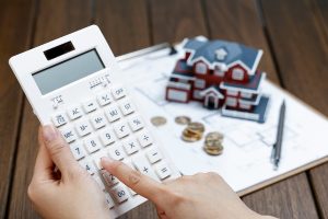 Main utilisant une calculatrice avec une maquette de maison et des pièces de monnaie en arrière-plan.