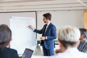 Un homme en costume fait une présentation devant un tableau blanc et plusieurs personnes assistent à la formation