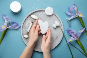 Mains tenant une pierre gua sha blanche sur une assiette grise avec des outils de massage du visage et des fleurs violettes sur un fond bleu