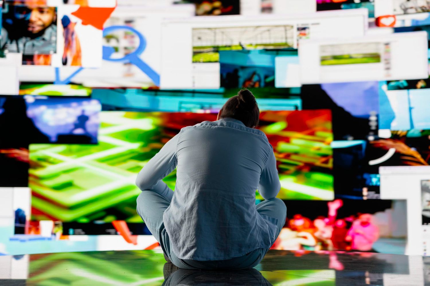 Une personne assise de dos face à un mur d'écrans affichant une multitude de contenus vidéo et d'images colorées simultanément.