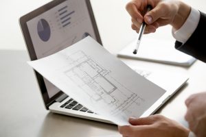 Expert analysant un plan architectural devant des graphiques financiers.