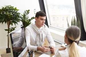Un jeune homme en chemise blanche discute sérieusement avec une thérapeute blonde vue de dos à un bureau équipé d'un ordinateur.