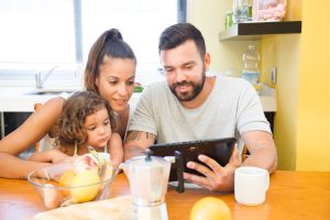 Une famille composée d'un père, d'une mère et d'une petite fille est réunie autour de la table de la cuisine. Le père tient une tablette numérique qu'ils regardent tous les trois ensemble
