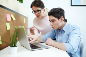 Une femme portant des lunettes se tient debout à côté d'un homme assis au bureau et montre du doigt l'écran de son ordinateur portable
