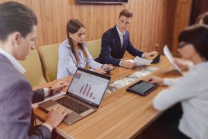 Une réunion de travail autour d'une grande table en bois. Plusieurs professionnels collaborent à l'aide d'ordinateurs, de tablettes et de documents papier