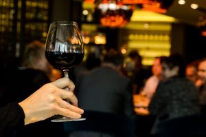 une main tenant un verre de vin rouge au premier plan dans l'ambiance feutrée et chaleureuse d'un restaurant ou d'un bar élégant.