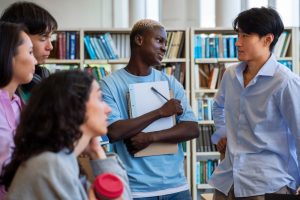 un groupe d'étudiants d'origines diverses discutant ensemble au milieu d'une bibliothèque garnie d'étagères de livres