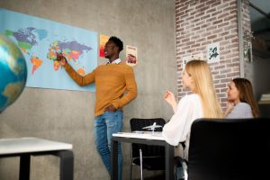un enseignant pointant un endroit sur une carte du monde affichée au mur, tout en s'adressant à des étudiantes installées à un bureau