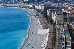 une vue aérienne panoramique de la baie des Anges et de la Promenade des Anglais à Nice, avec la plage de galets bordée par une mer bleue turquoise et les bâtiments de la ville.