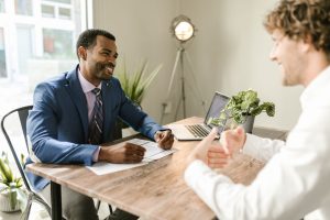 conseiller en patrimoine souriant, en costume bleu et cravate, échangeant à un bureau en bois avec un client assis en face de lui