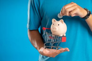une pièce dans une tirelire en forme de cochon, placée sur un mini chariot de supermarché tenu dans la main
