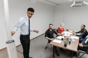 Un professionnel présente une démonstration sur tableau à ses collègues attentifs réuni·e·s autour d'une table de réunion dans un bureau moderne.