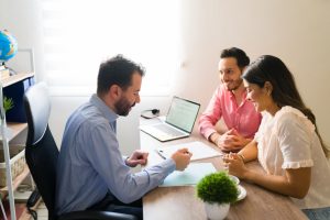 Un conseiller professionnel en chemise bleue échange avec un couple souriant assis en face de lui autour d'un bureau de rendez-vous.