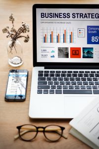 un ordinateur portable affichant des données de stratégie d'entreprise, entouré d'un smartphone, de lunettes et d'un vase décoratif sur un bureau en bois