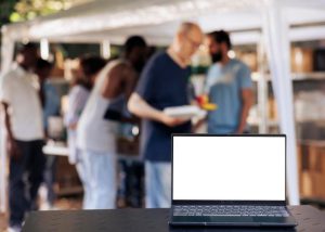 Un ordinateur portable à écran blanc ouvert au premier plan, posé sur une table devant un groupe de personnes floues rassemblées en extérieur
