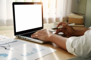 une personne en train de travailler sur un ordinateur portable, tenant un stylo tout en tapant au clavier sur un bureau agrémenté de documents imprimés et de graphiques