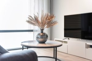 Un salon moderne et épuré avec un vase décoratif garni d'herbes de pampa posé sur une table basse ronde, à côté d'un meuble TV blanc supportant un grand écran plat près d'une fenêtre lumineuse.