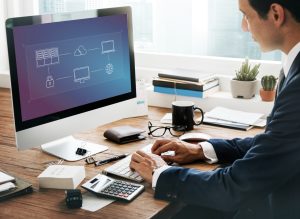 Un homme d'affaires en costume travaille à son bureau face à un grand écran d'ordinateur affichant un schéma d'architecture réseau et cloud