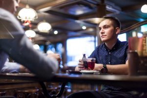 Un jeune homme en chemise sombre à motifs écoute attentivement son interlocuteur assis en face de lui autour d'une boisson